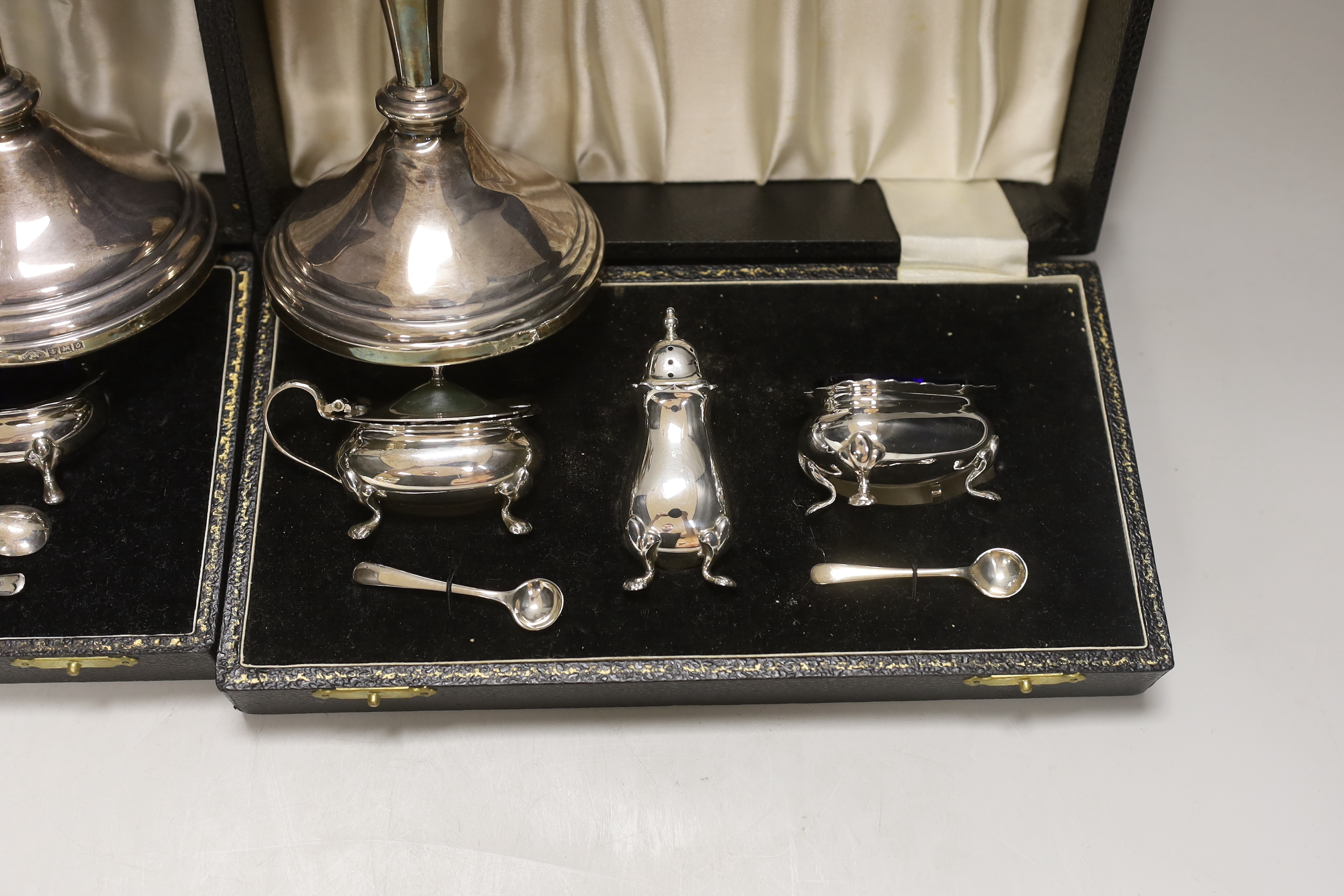 Two modern cased silver three piece cruet sets, with spoons and a pair of modern silver candlesticks, 19.5cm, weighted.